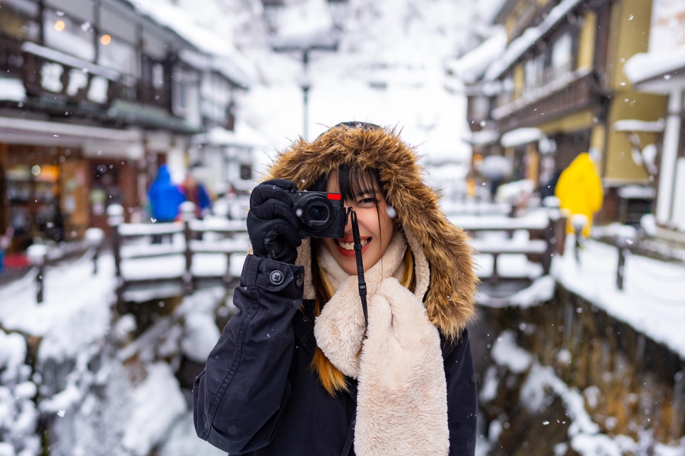 Woman holding a camera to her face wearing a hooded coat and fluffy scarf, one of the best outfits in Japan for Winter. In the background is a snowy, small town in Japan.