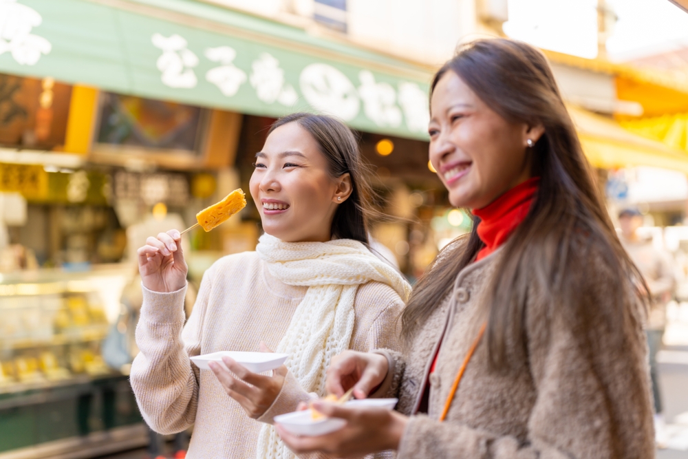 Two young woman in sweaters holding street food at a market and showing off what to wear in Japan in the fall.