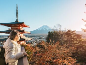 Woman in a coat showcasing what to wear in Japan in the fall stands looking out over fall foliage near a pagoda and Mount Fuji in the distance.