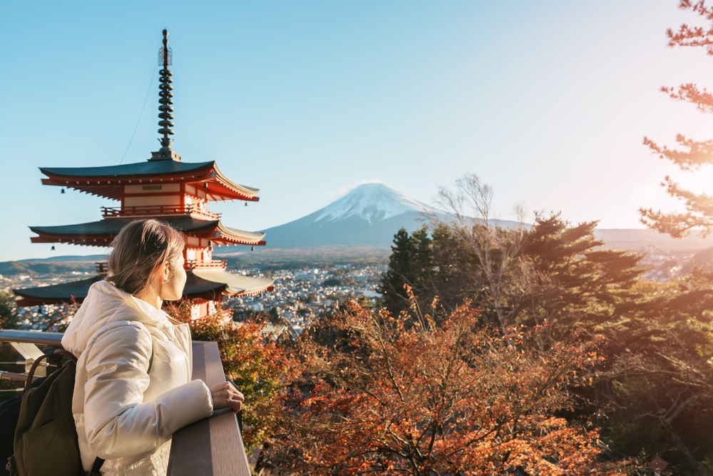 Woman in a coat showcasing what to wear in Japan in the fall stands looking out over fall foliage near a pagoda and Mount Fuji in the distance.