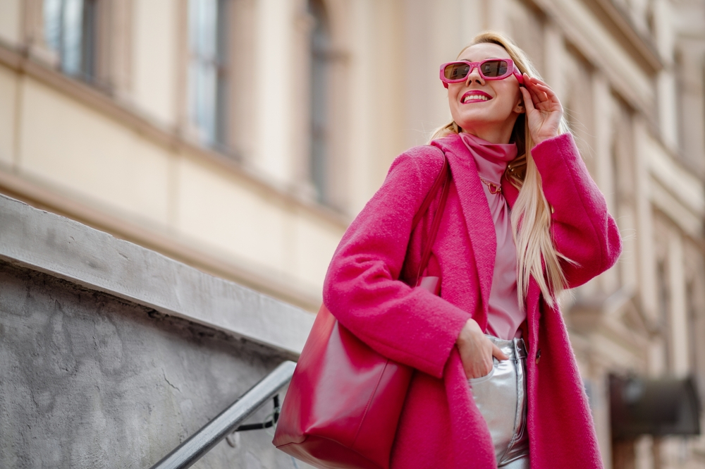 Woman wearing fuchsia color coat, sunglasses, pink turtleneck, holding faux leather tote, shopper bag, posing in street of city. 