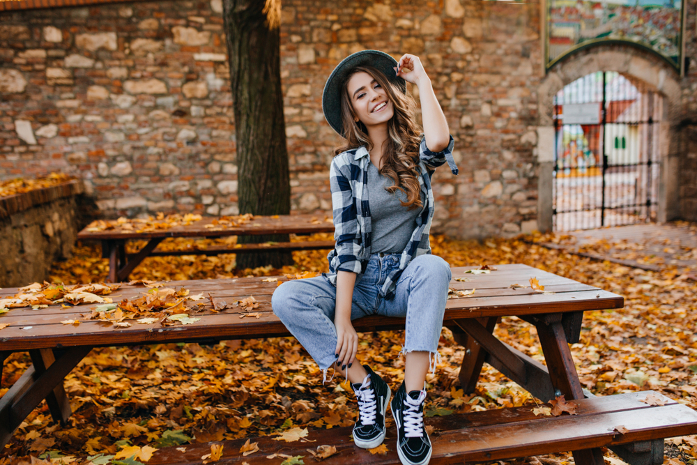 Carefree young woman in trendy vintage pants sitting on table in park and laughing. The article is about London in fall. 