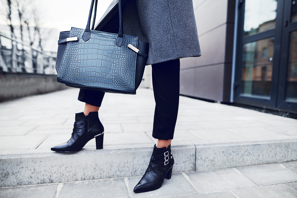 Fashionable woman posing in street, holding big dark blue textured bag, wearing stylish pointed toe ankle boots. The article is about London in fall. 