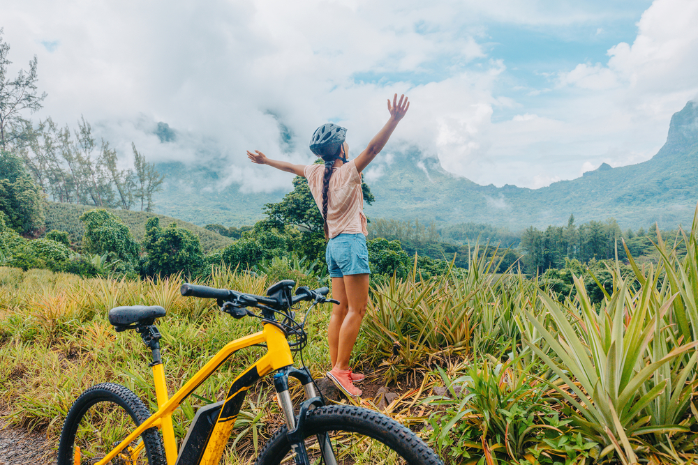 Woman standing with her hands in the air next to a mountain bike in the jungle of Tahiti on a cloudy day.