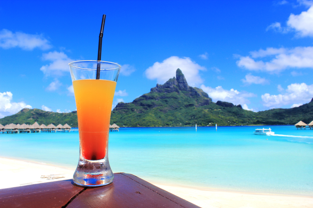Orange colored cocktail in front of a background of the bright blue ocean and mountain.