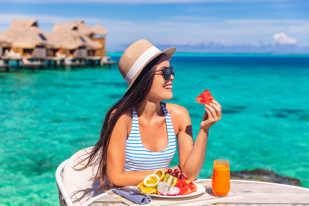 Woman in sunglasses and wearing a hat while eating a plate of fruit in front of the ocean.