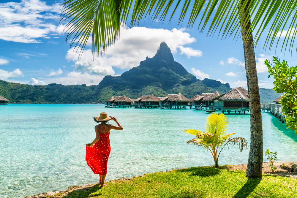 Woman in a long red dress and sun hat stands under a palm tree overlooking the ocean with over water bungalows and a pointed mountain in the distance.