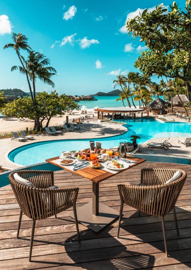 A table set with a tasty meal overlooking a pool among palm trees with the ocean in the distance at Le Bora Bora by Pearl Resorts.