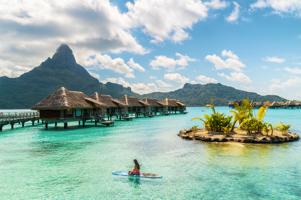 Wide-angle photo of a woman sitting on paddle board in the ocean near over water bungalows.