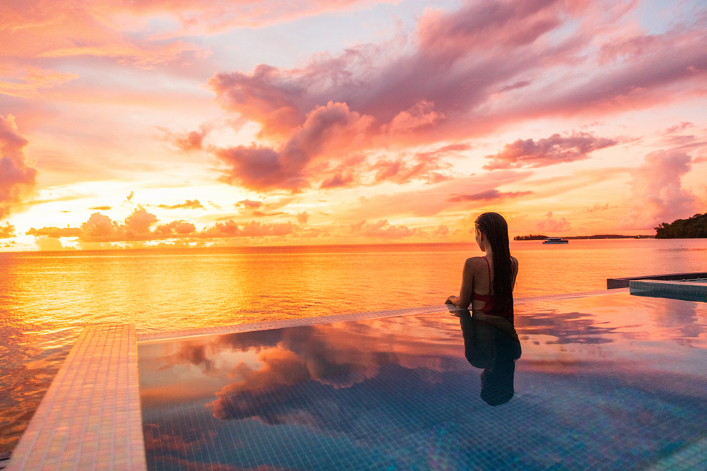 Woman in a pool overlooking a bright, warm sunset over the ocean.