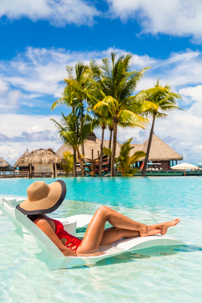 Woman in a red swim suit and sun hat sits in a lounge chair in shallow water with bungalows in the background.