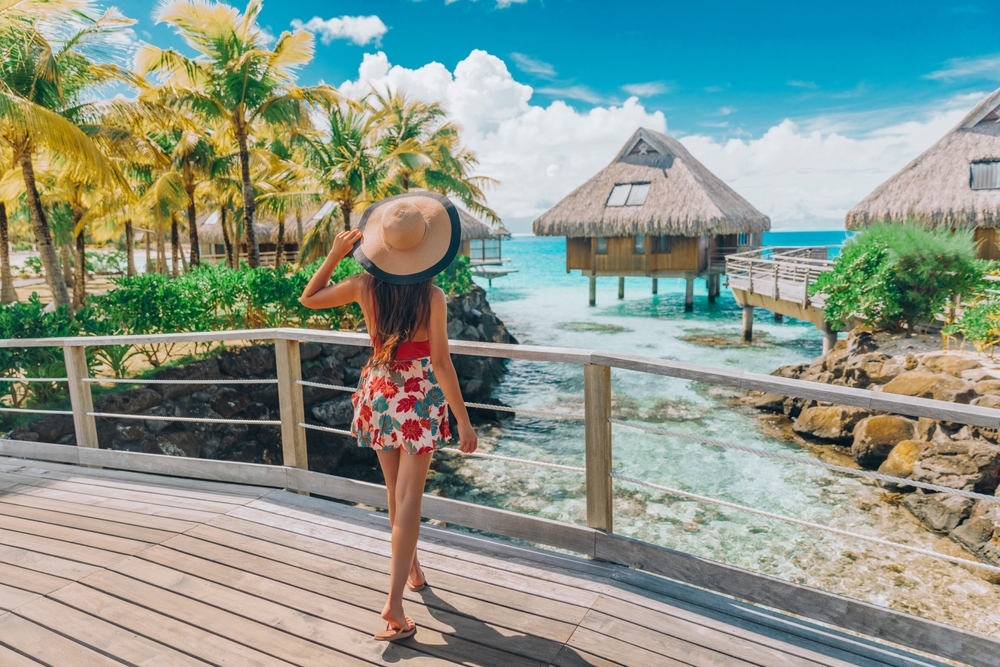 Woman in a dress and sun hat stands on a bridge looking out at the ocean and bungalows.