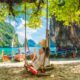 Woman in a hat sitting on a swing on Lao Lading beach with traditional boats in the water while traveling to Thailand.
