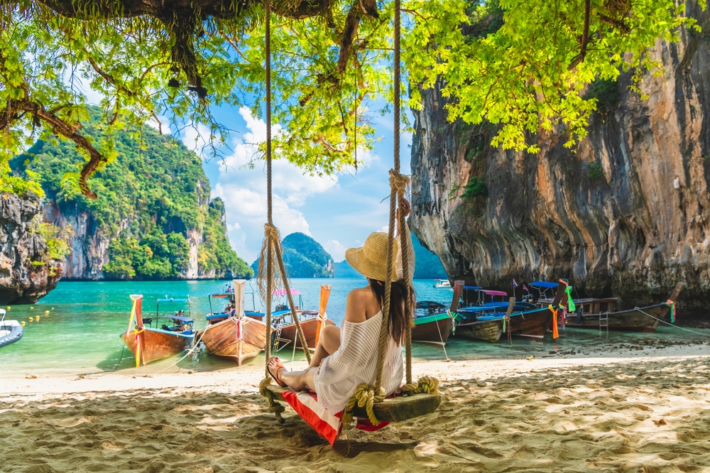Woman in a hat sitting on a swing on Lao Lading beach with traditional boats in the water.