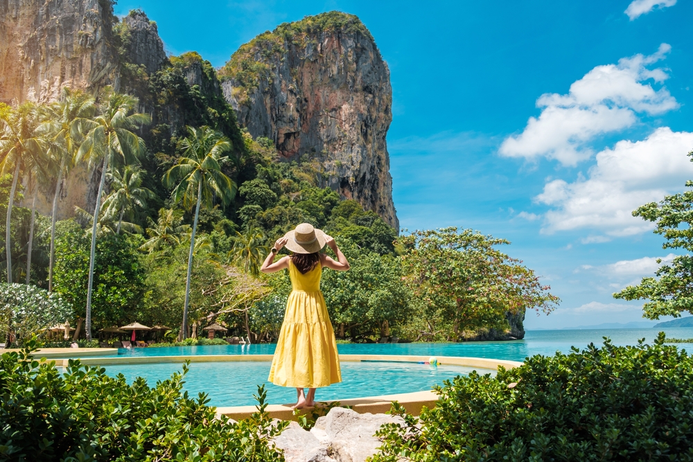 A woman in a long, yellow dress and a hat stands looking out at the water and lush forest in Thailand.