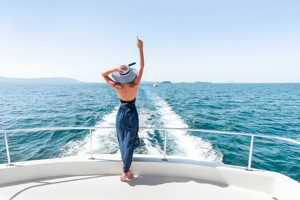 Rear,View:,A,Young,Girl,In,A,Blue,Dress,Is Rear view: a young girl in a blue dress is standing, raising her hands on the edge of a yacht and admiring the scenery of the azure sea. Travel and vacation