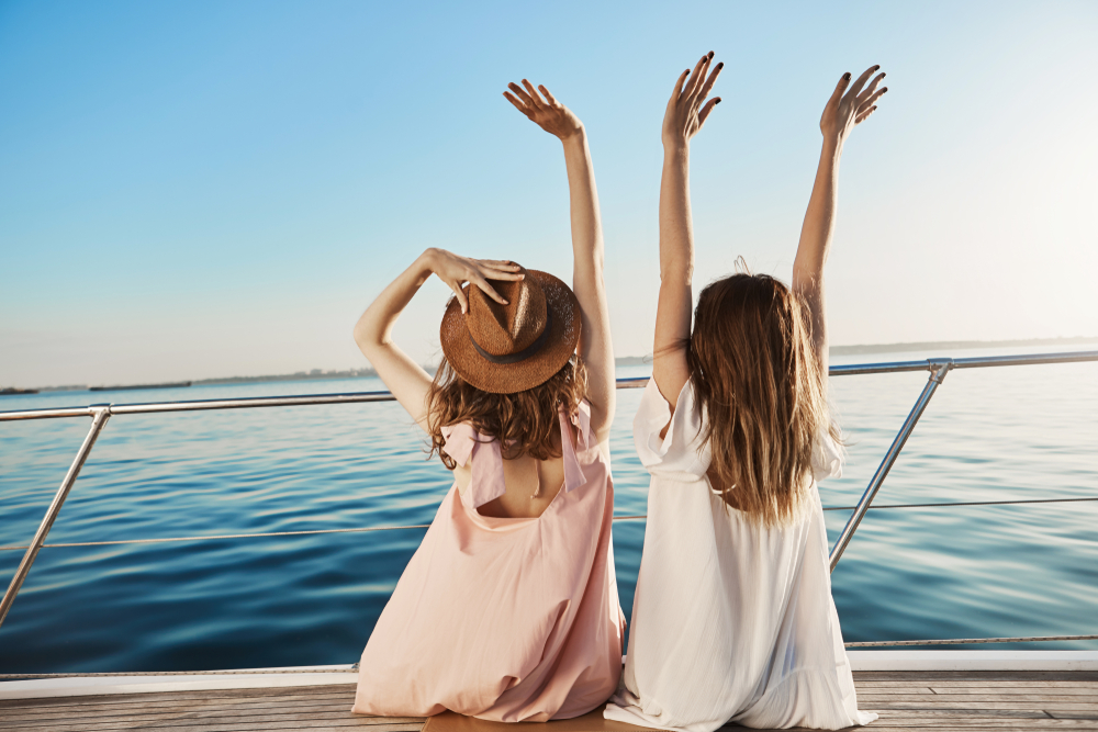 Two girls on a boat deck with their arms in the air. One is wearing a hat.