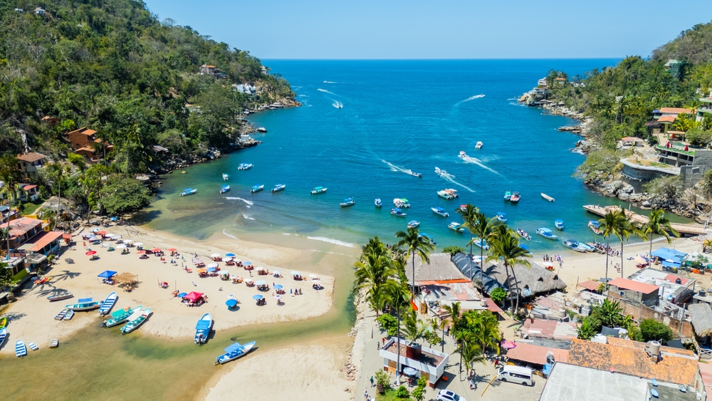 deep blue ocean water with green-treed mountains on either side and boats in water next to sandy beach.