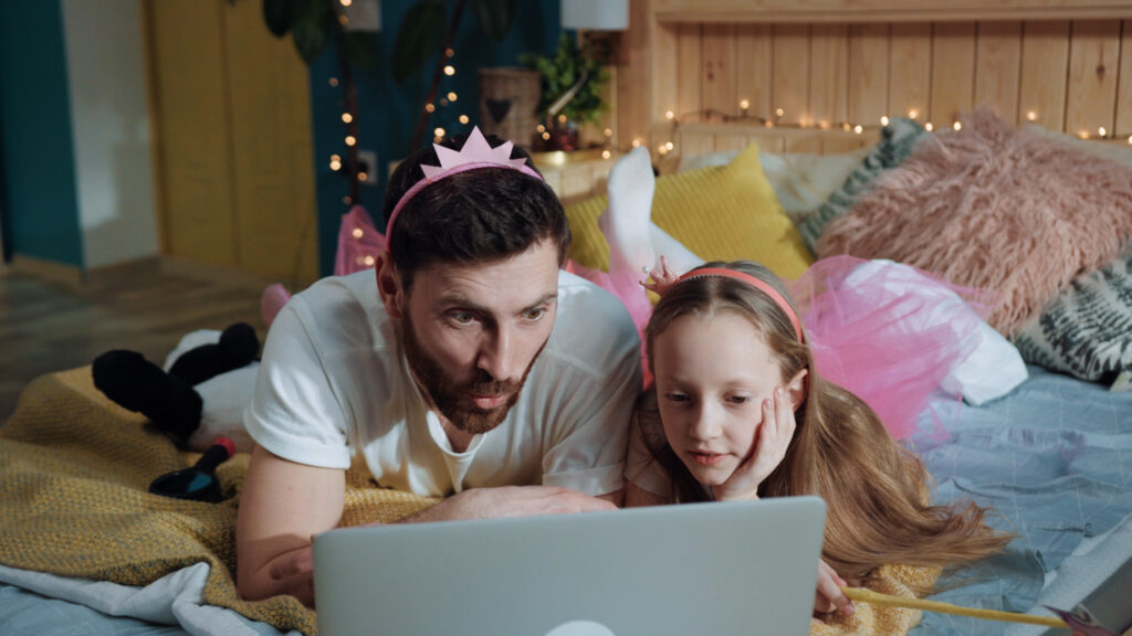 Dad and young daughter both wearing pink headbands laying on bed looking into computer.