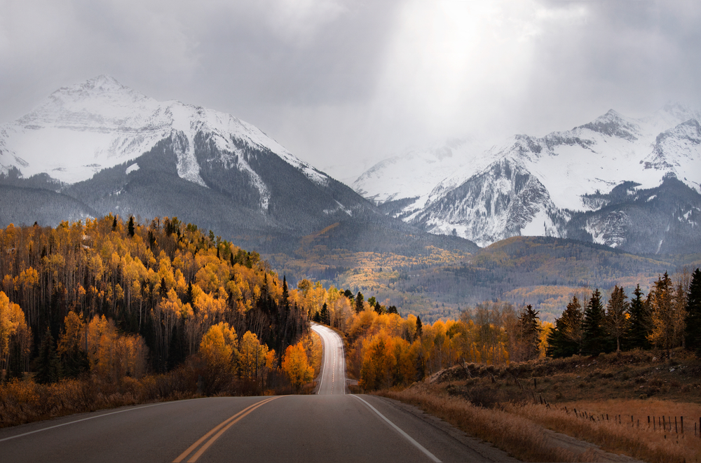 Light rays shine down on a scenic road in Colorado surrounded by mountains and fall colors. An autumn mountain scene near Mountain Village & Telluride. Light rays shine down on a scenic road in Colorado surrounded by mountains and fall colors. An autumn mountain scene near Mountain Village & Telluride. Light rays shine down on a scenic road in Colorado surrounded by mountains and fall colors. An autumn mountain scene near Mountain Village & Telluride. The article is about a Colorado road trip. 