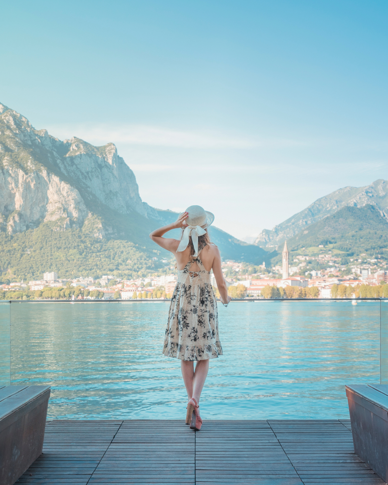 A girl with a hat looking at Lake Como and Lecco city from Malgrate in a sunny day. The article is about a Lake Como packing list. 