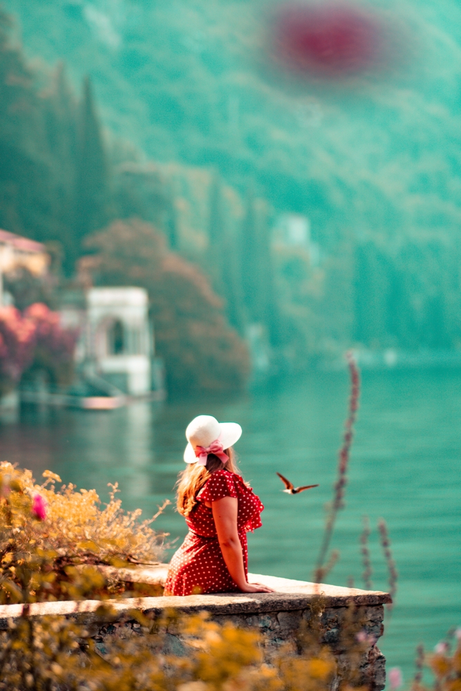 A woman in a red polka dot dress poses with a view of Lake Como from the botanical garden of Villa Monastero, Varenna, Italy. The article is about a Lake Como packing list. 