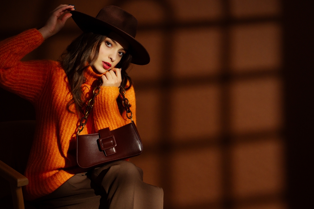 Fashionable confident beautiful brunette woman wearing trendy hat, orange sweater, holding stylish leather purse, posing on brown background. 