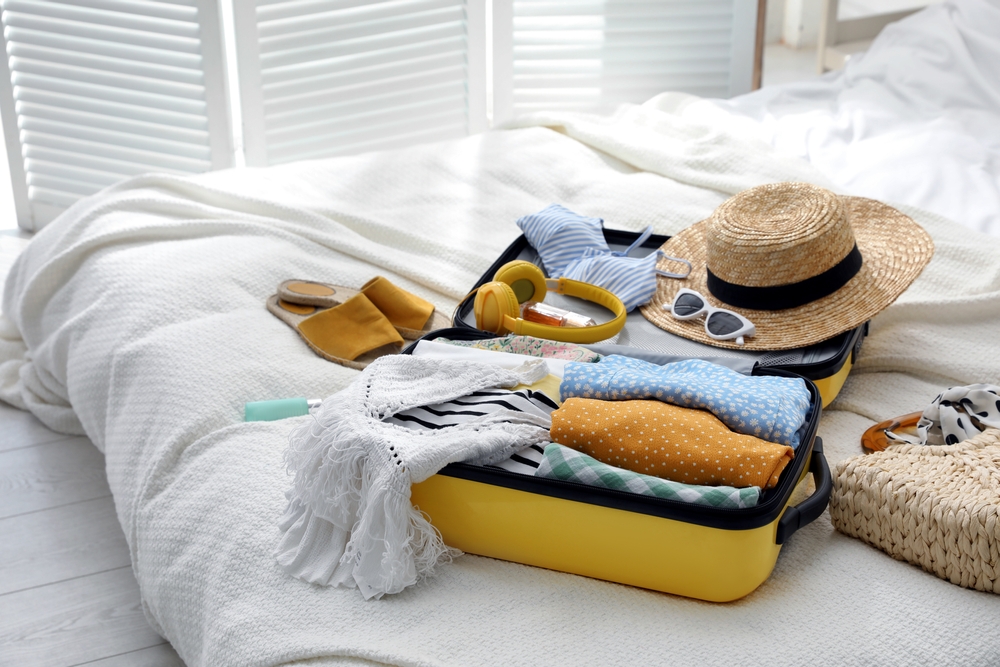 Open suitcase full of clothes, shoes and summer accessories on bed. The article is about what to pack for a Mediterranean cruise.