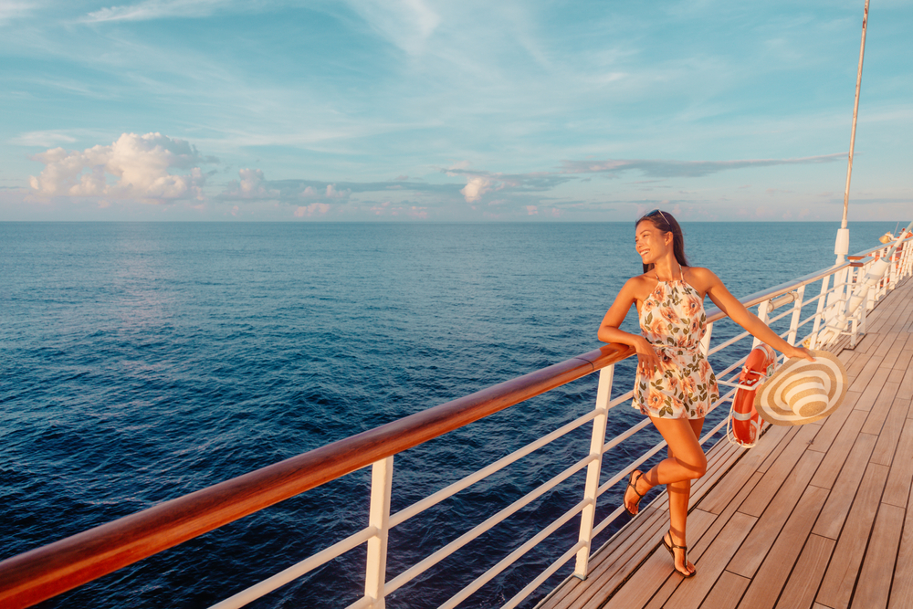 Tourist woman watching sunset from balcony deck. She has a cute dress on and a hat. The article is about what to pack for a Mediterranean cruise.