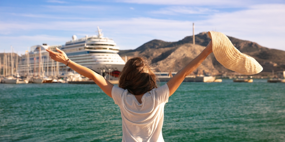 Woman looking at boat in the sea with her arms out stretched and a hat in her hand.
