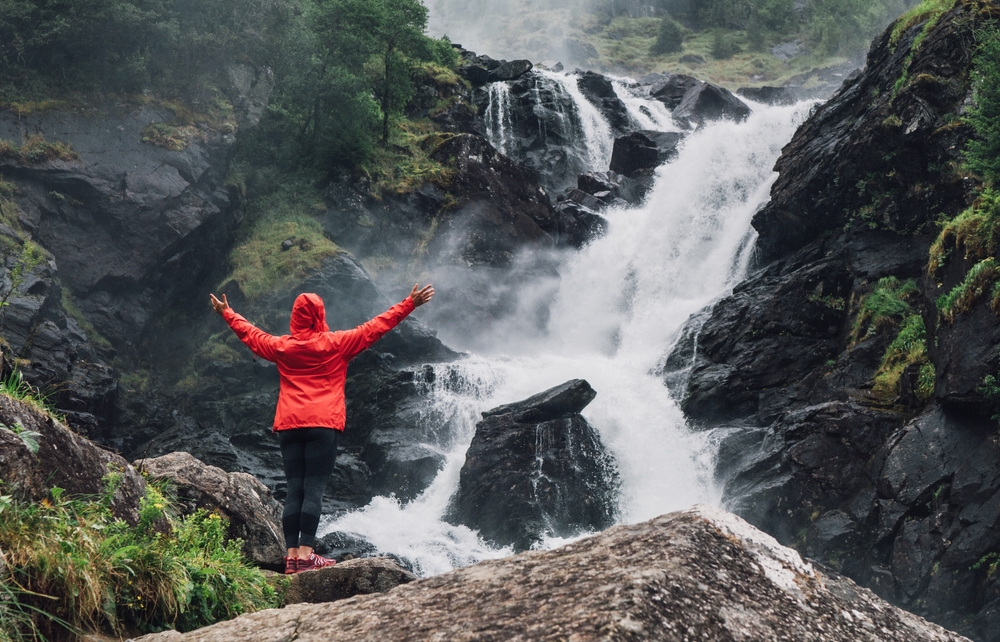 woman in red jacket standing in front of a waterfall traveling to norway