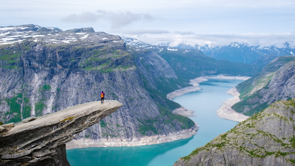 a person standing on the edge of a cliff overlooking a river surrounded by mountains traveling to norway