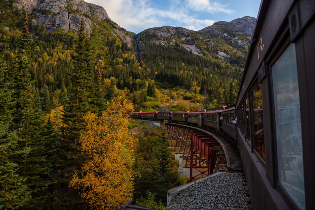 Old Historic Railroad Train is going over a wooden bridge up White Pass with tourists during a cloudy summer morning. The article is about train trips in the United States.
