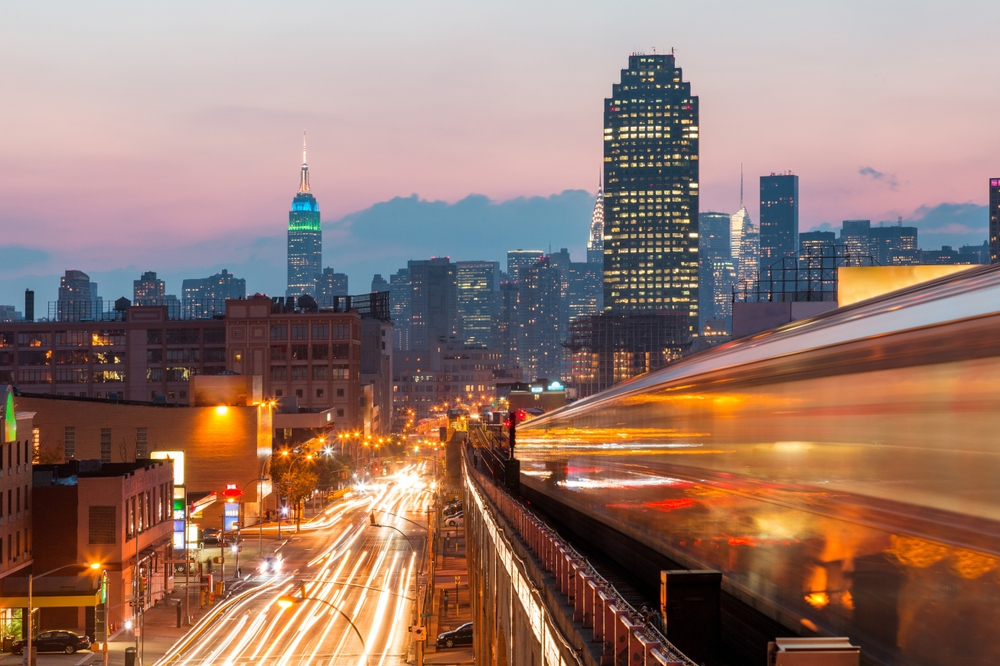 New York view at sunset with blurred train and busy roads on foreground and Manhattan skyline on background. The article is about train trips in the USA