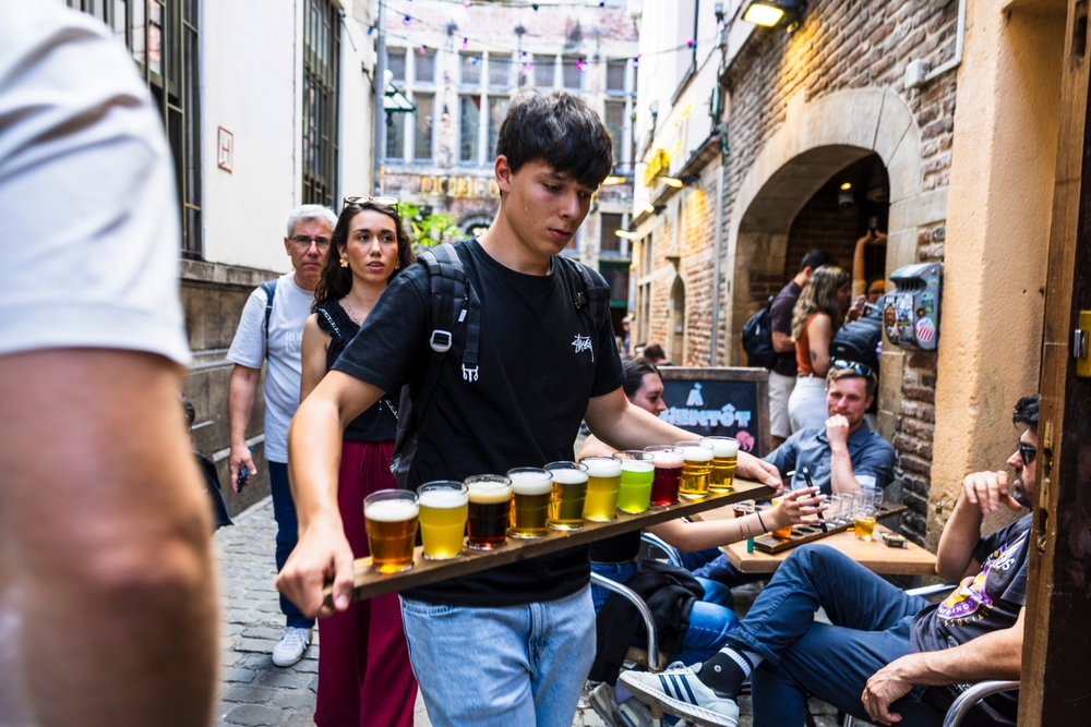 A young man carries a long tray filled with glasses of different beers through a busy alley bar in Brussels.