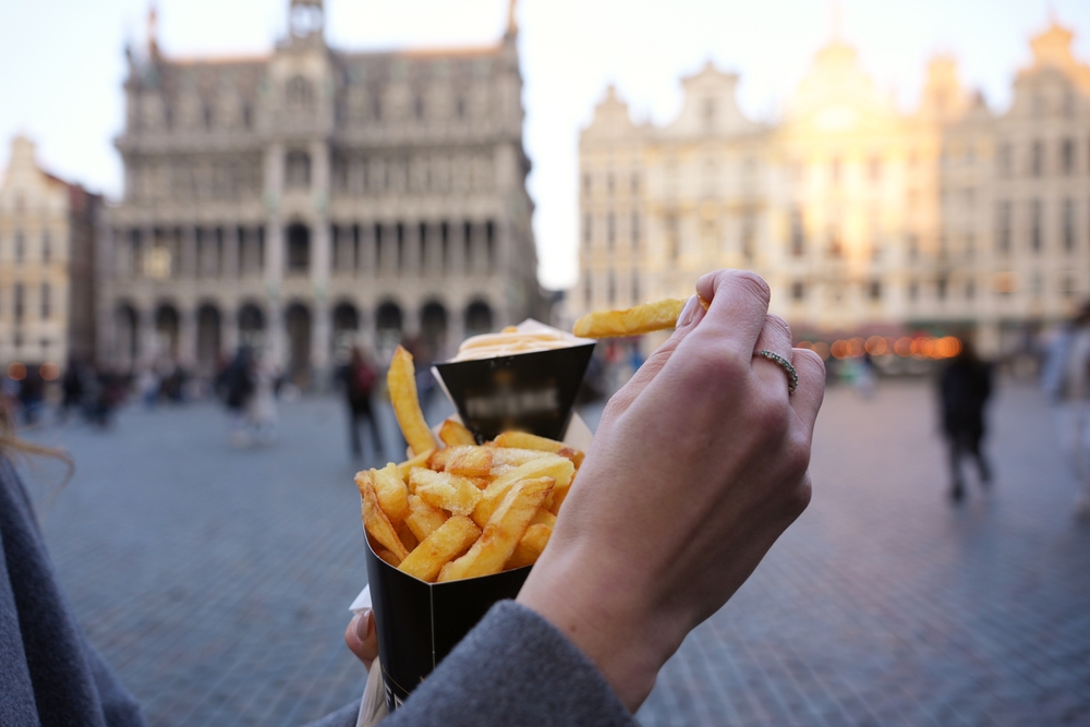 Close-up of a hand holding classic Belgian fries with sauce in Grand Place, Brussels, with golden light on historic buildings in the background.