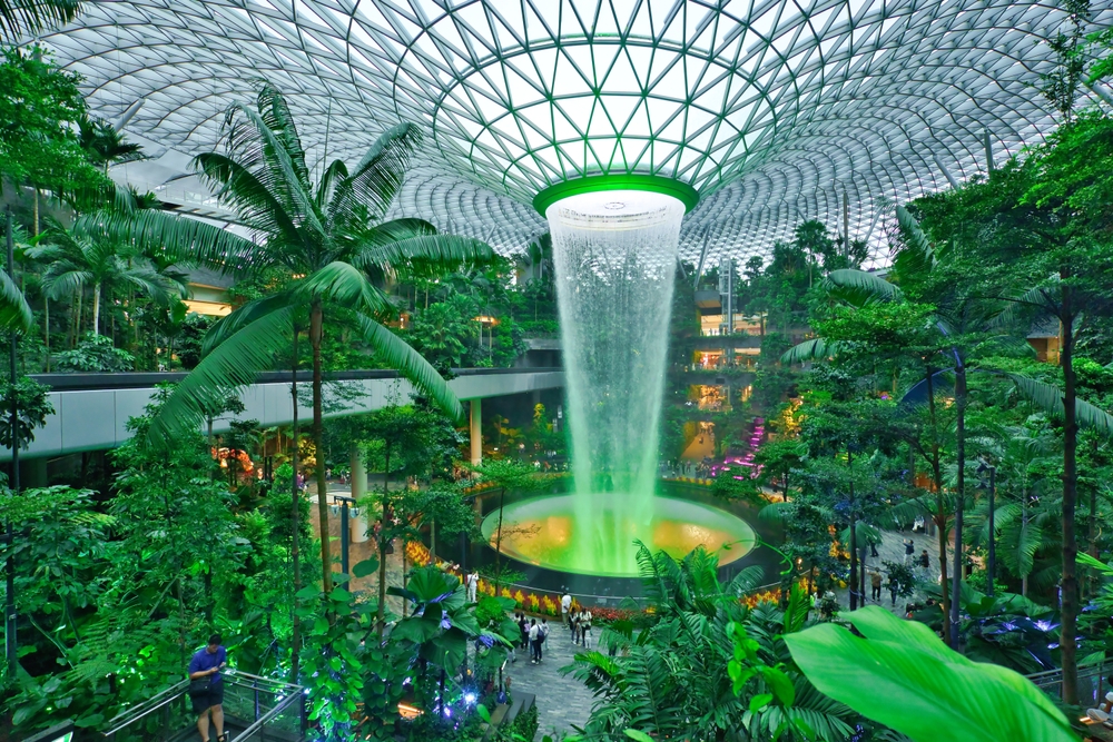 large waterfall in middle of photo with intricate glass ceiling surrounded by green foliage.