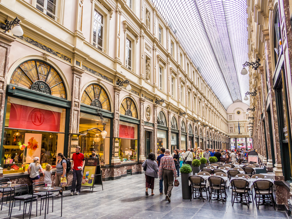 People are walking and there are tables and chairs inside fashionable shopping area.