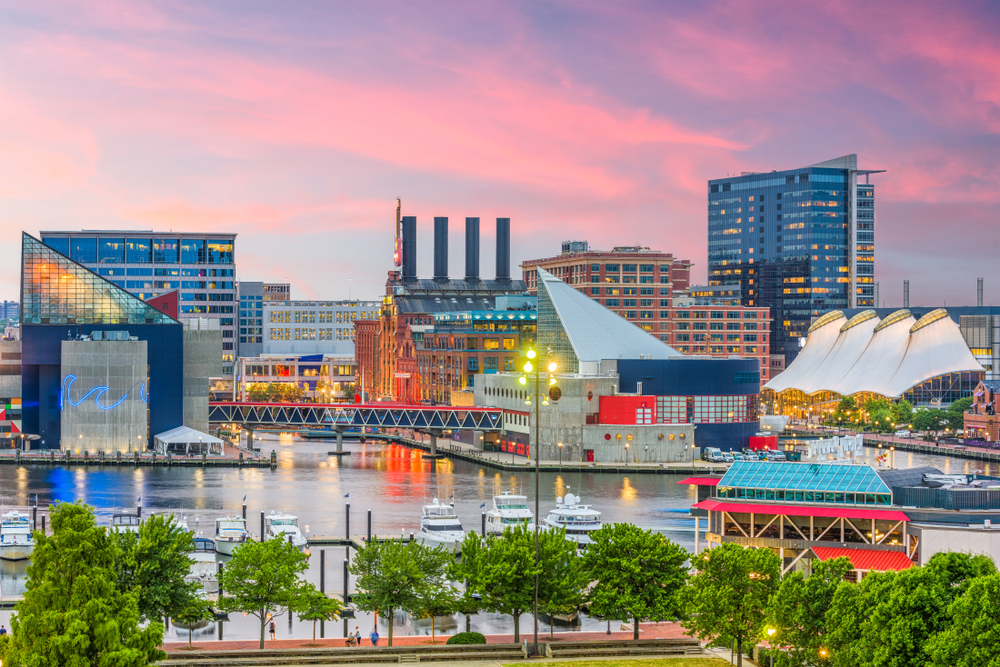 Skyline over the Inner Harbor at dusk. There are cit building in the background and trees in the foreground. The article is about a Baltimore weekend getaway.